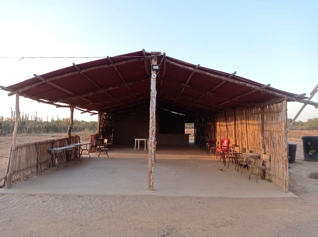 A completed open-air community building with cane walls, a red metal roof, a concrete floor, desks and chairs.