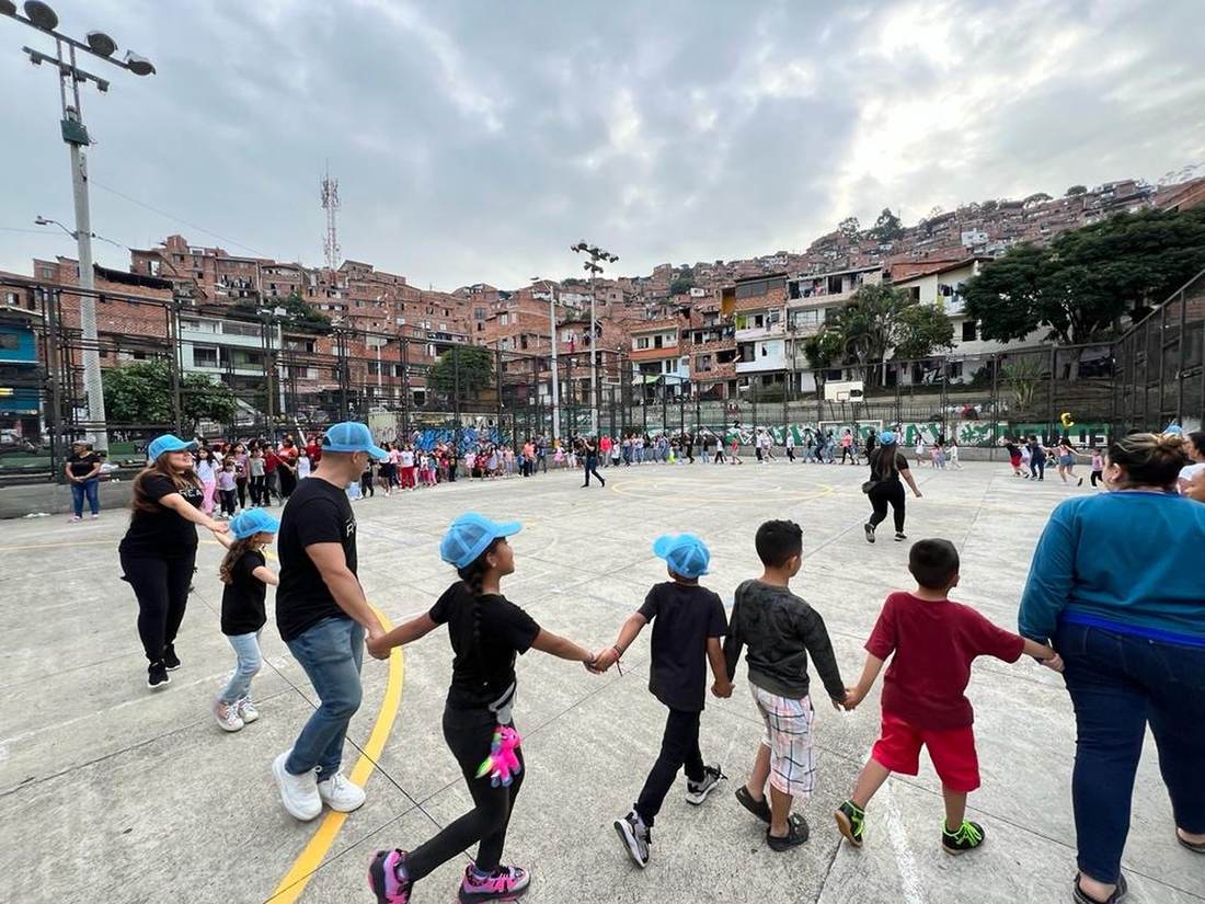 Children and volunteers in matching caps walking hand in hand in a circle on a concrete court, with a hillside neighbourhood behind them.