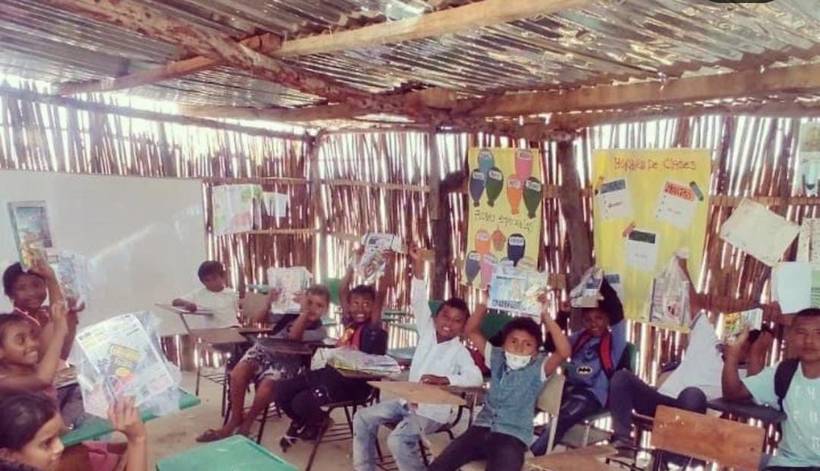 Children at desks in an open-air classroom with cane walls, holding up their workbooks, with class-schedule posters on the wall behind them.