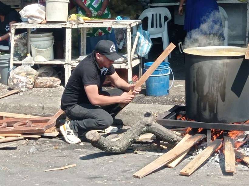 A Fundación Cloud volunteer crouching to feed split wood into an open fire beneath a large steaming cooking pot in a street.