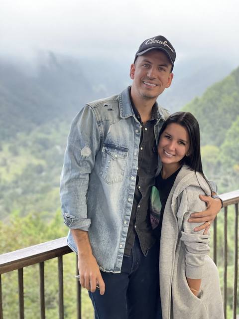 Joel and Giselle Manderfield standing together on a balcony with forested mountains behind them.