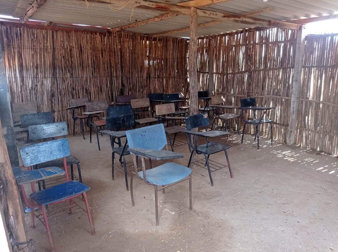 The empty interior of a cane-walled classroom with a corrugated roof and about a dozen mismatched second-hand school desks.