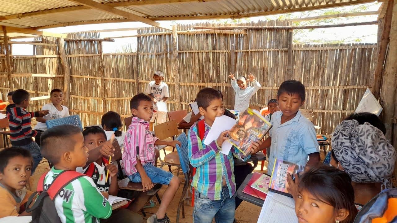 Children at desks in an open-air classroom with cane walls, holding up their workbooks, with class-schedule posters on the wall behind them.