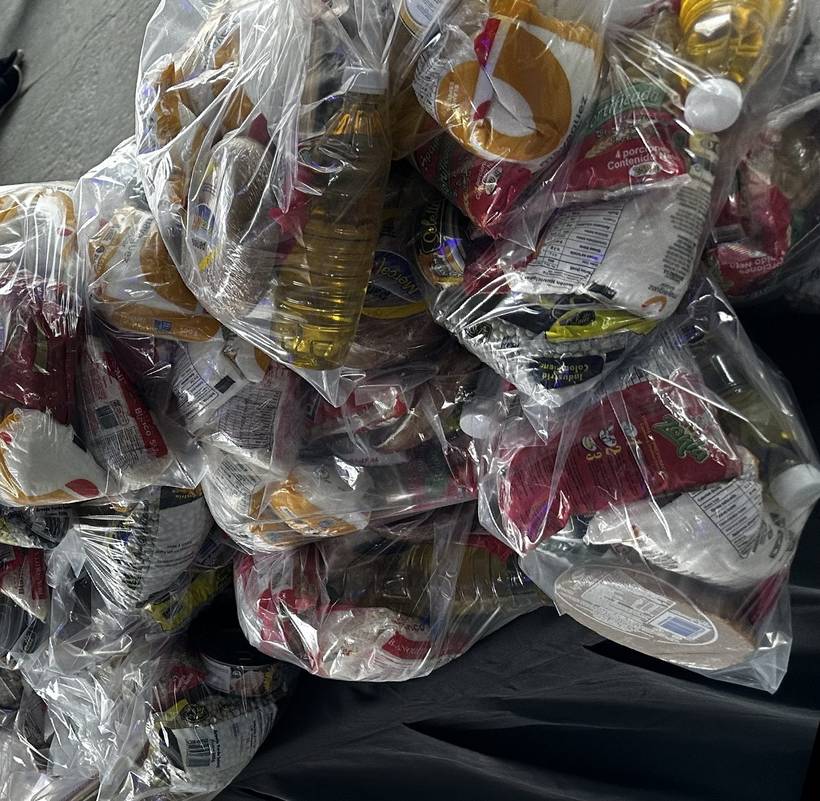 Dozens of clear family food packages bagged and stacked on a table, containing cooking oil, rice, pasta, beans and panela.