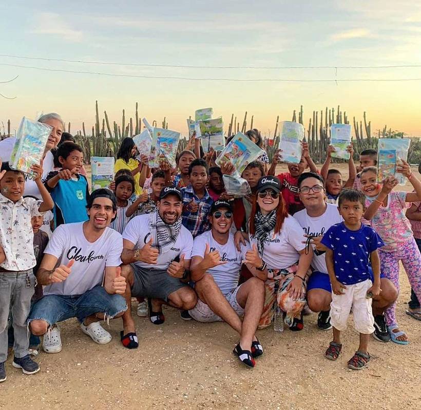 Children holding up packs of school supplies above their heads at sunset in La Guajira, with the Cloud team crouched in front.