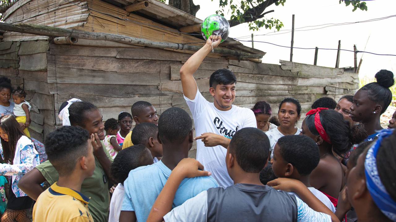 A Fundación Cloud volunteer in a white shirt holds up a ball, surrounded by a crowd of children and teenagers.