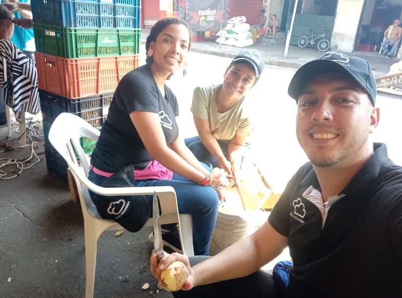 Three Fundación Cloud volunteers sitting on plastic chairs peeling potatoes at a market, smiling at the camera.